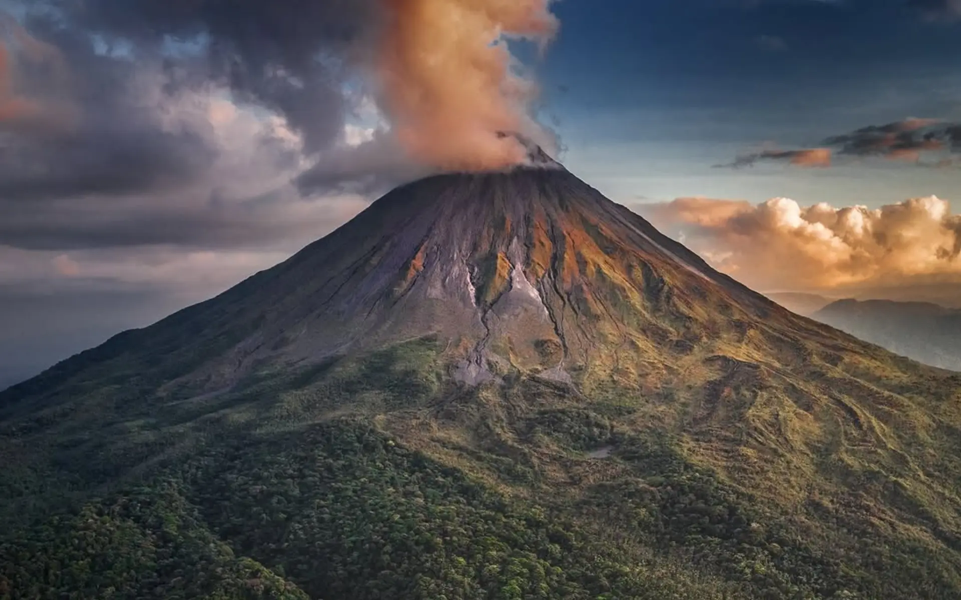 Arenal Volcano with a plume of orange-lit smoke at its summit above green forested slopes