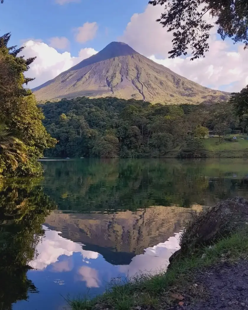 Volcano reflected in a calm lagoon, with forest along the far shore under a blue sky