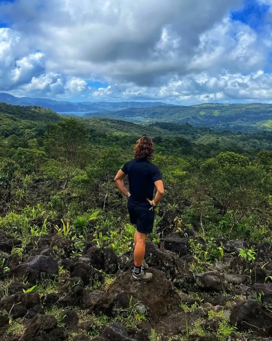 Hiker in dark clothes standing on black lava rocks, looking out over forested hills and a distant lagoon