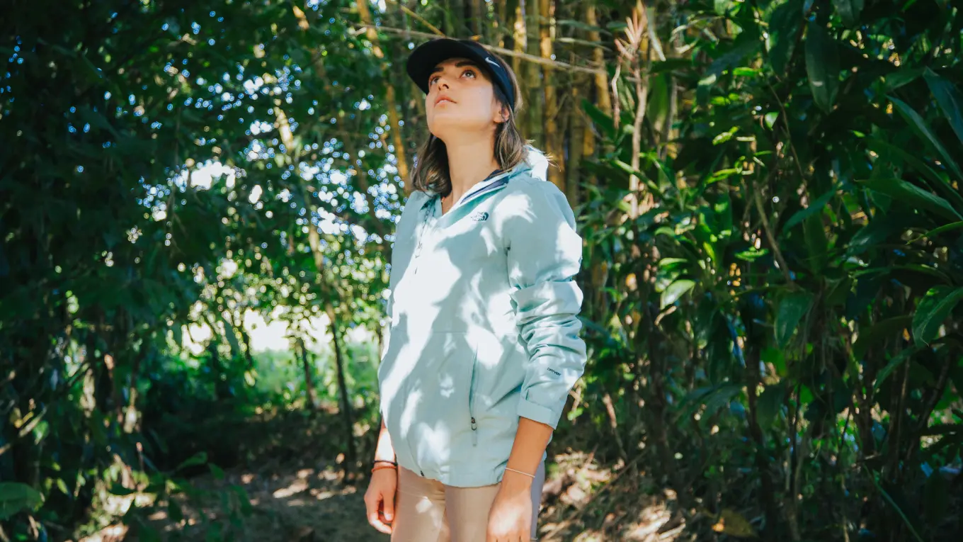 Woman in a cap and a light jacket looking up into the trees on a shaded forest path
