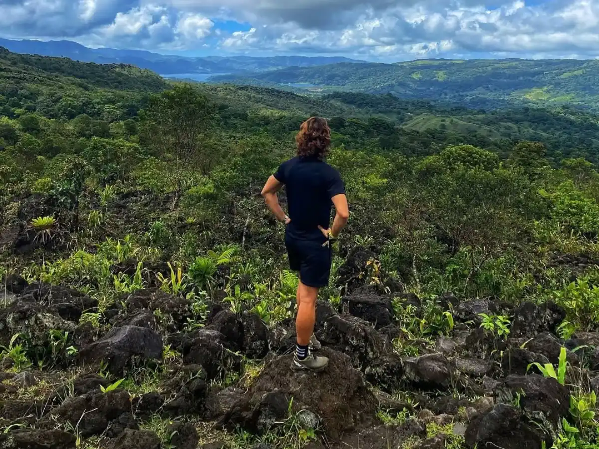 Hiker with hands on hips standing on dark lava rocks, looking over green forest and distant hills
