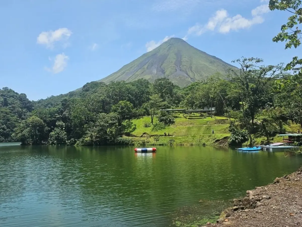 Green lagoon with a floating trampoline, a lawn and forest, and the cone of Arenal Volcano behind