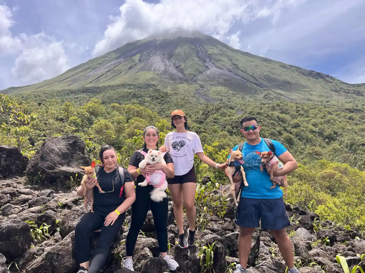 Four visitors holding small dogs on lava rocks, with Arenal Volcano under a cloud behind them