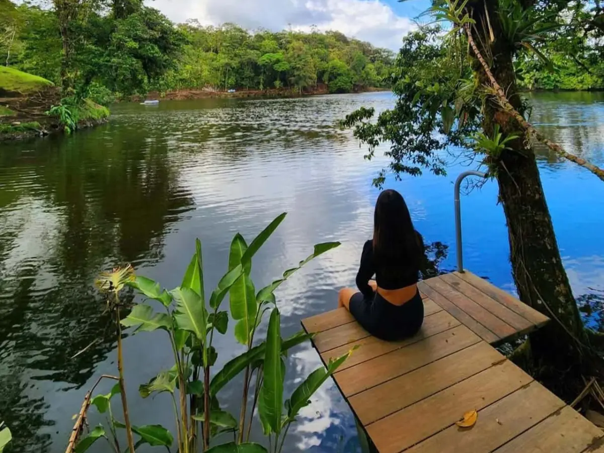 Woman seated on a wooden dock at the edge of a calm green lagoon, with banana leaves and forest around