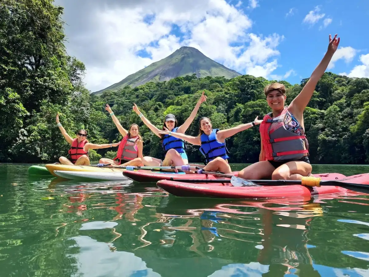 Cinco personas con chaleco salvavidas sobre tablas de paddle, con los brazos en alto y el Volcán Arenal al fondo