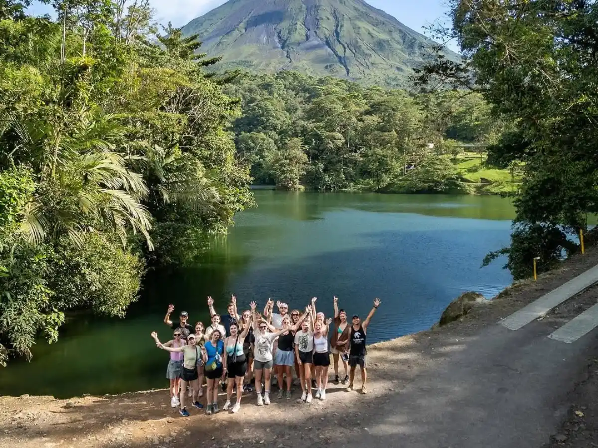 Grupo de visitantes con los brazos en alto a la orilla de la laguna, con el Volcán Arenal sobre el bosque