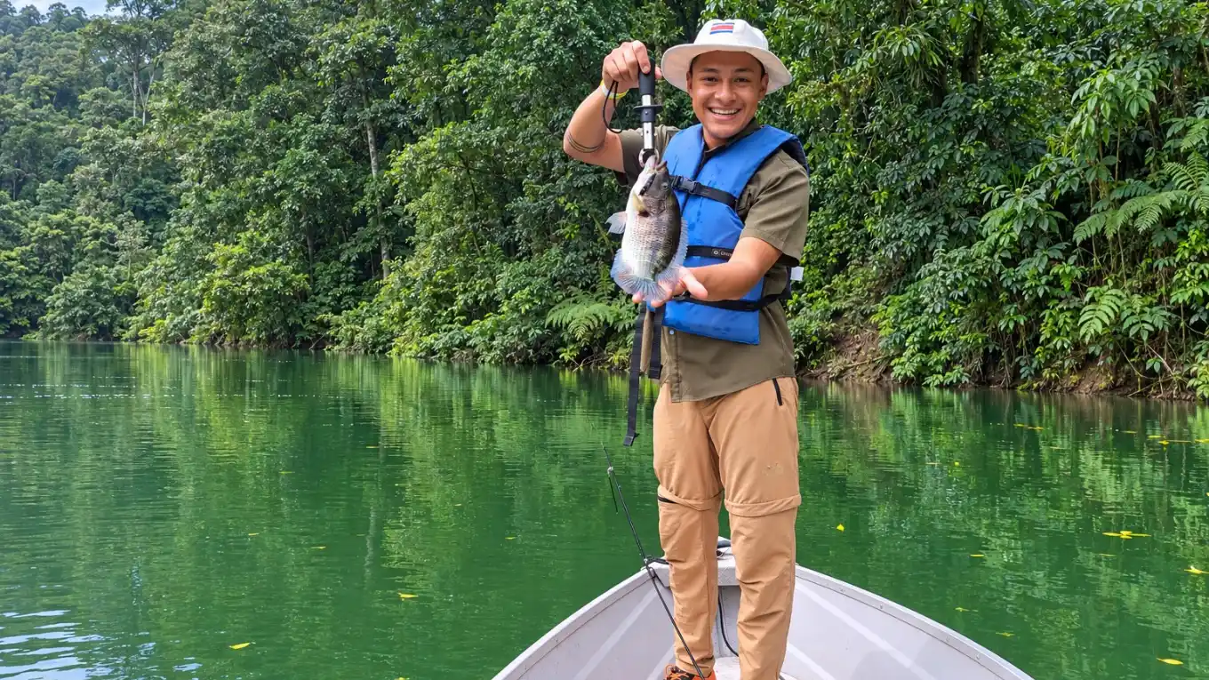 Smiling man in a hat and life jacket on a boat bow holding up a small fish, on a green forest-lined lake