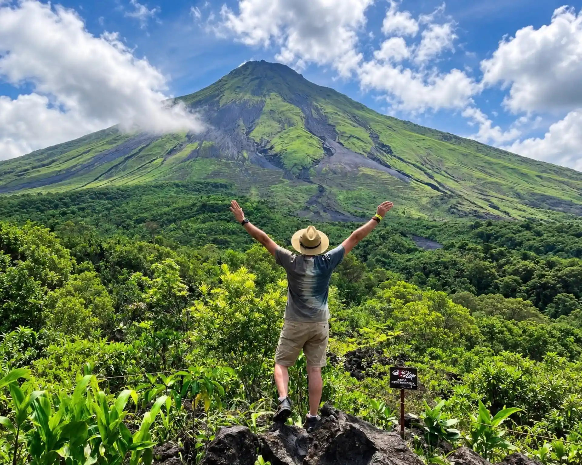 Hiker in a straw hat with arms raised on a lava rock, facing Arenal Volcano above a green forest