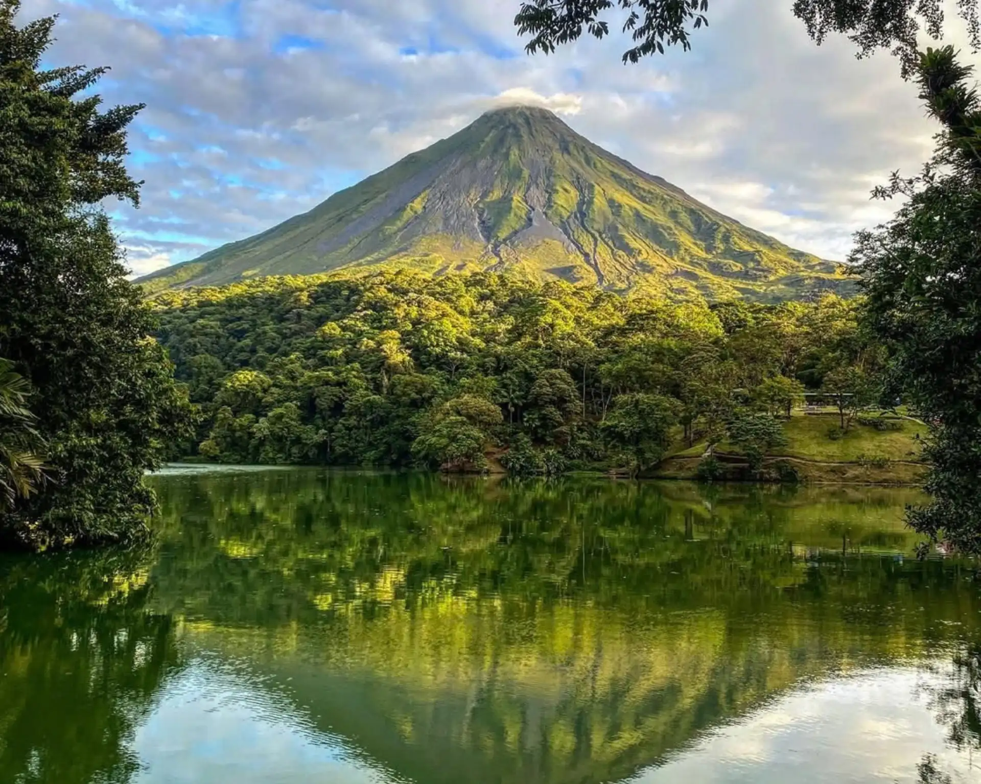 Arenal Volcano under soft clouds, framed by trees and mirrored in a still green lake