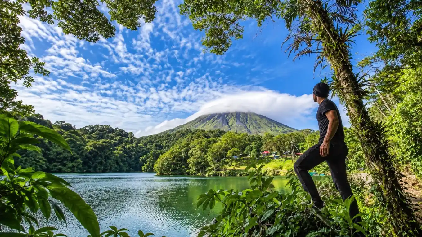 Man standing at a lagoon shore among tall trees, looking at a volcano with a cloud over its summit