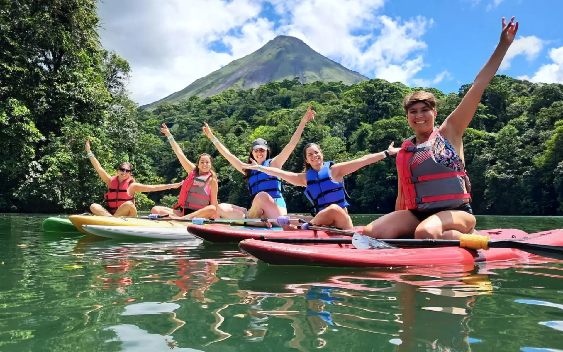 Cinco personas con los brazos en alto sobre kayaks y tablas de remo en una laguna, con el volcán Arenal al fondo
