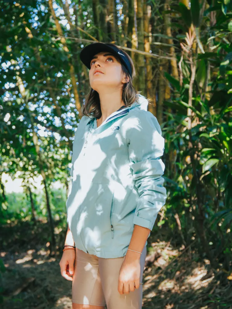 Persona con gorra y chaqueta clara que mira hacia arriba en un sendero del bosque, entre bambú y hojas