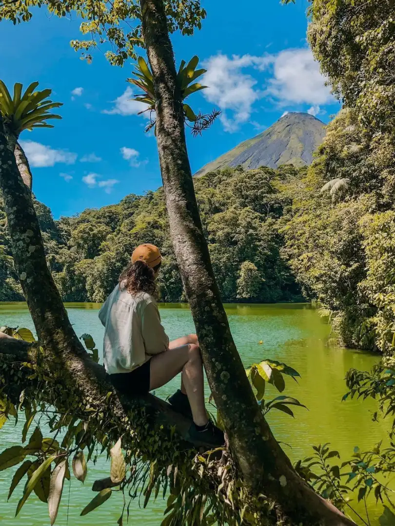 Persona con gorra naranja sentada en un tronco inclinado sobre una laguna verde, con un volcán al fondo
