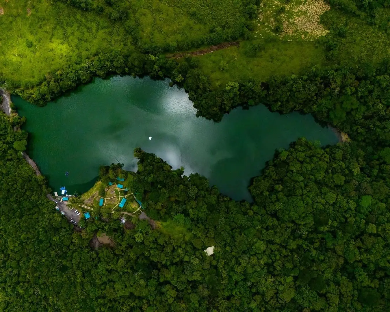 Vista aérea de una laguna verde rodeada de bosque denso, con un pequeño campamento en la orilla