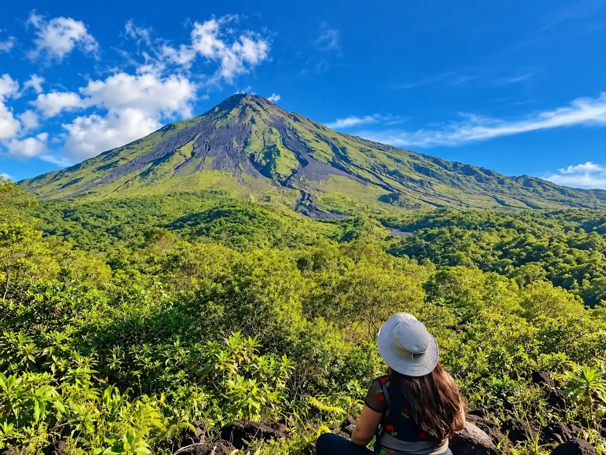 Persona con sombrero blanco sentada entre vegetación verde que mira hacia el volcán Arenal