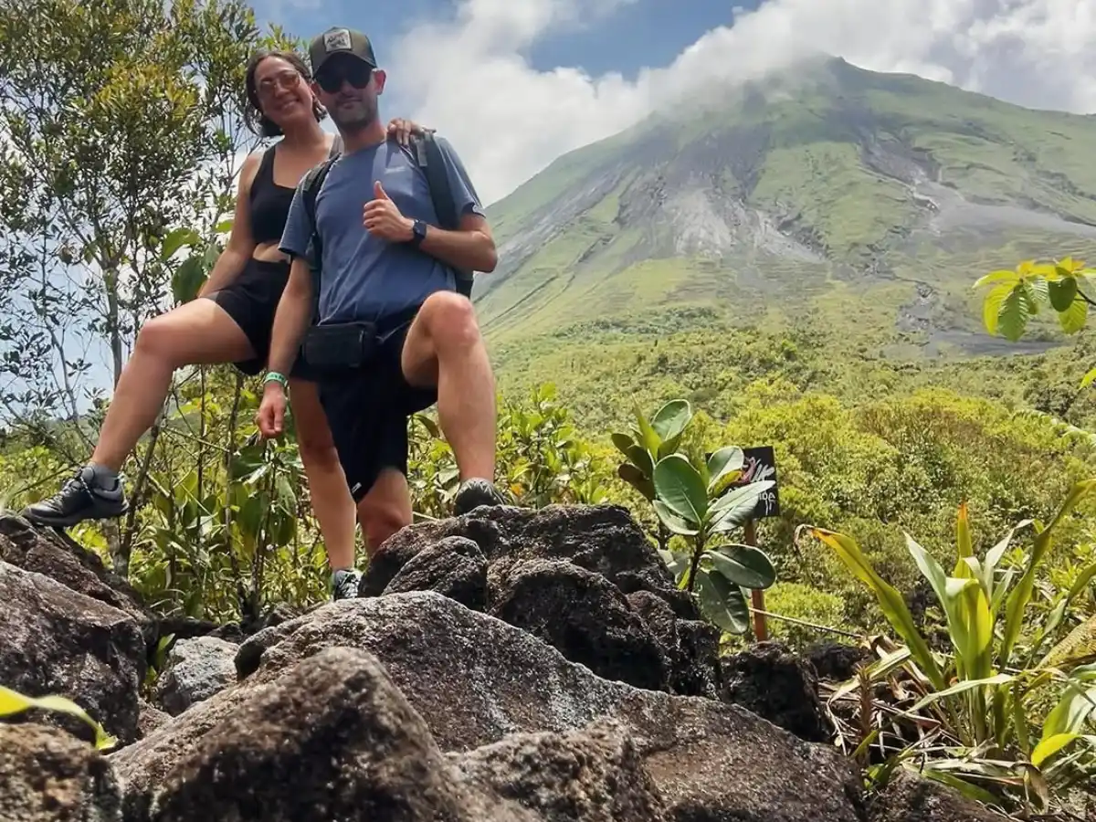 Dos personas posando sobre rocas oscuras con la ladera verde del volcán Arenal detrás