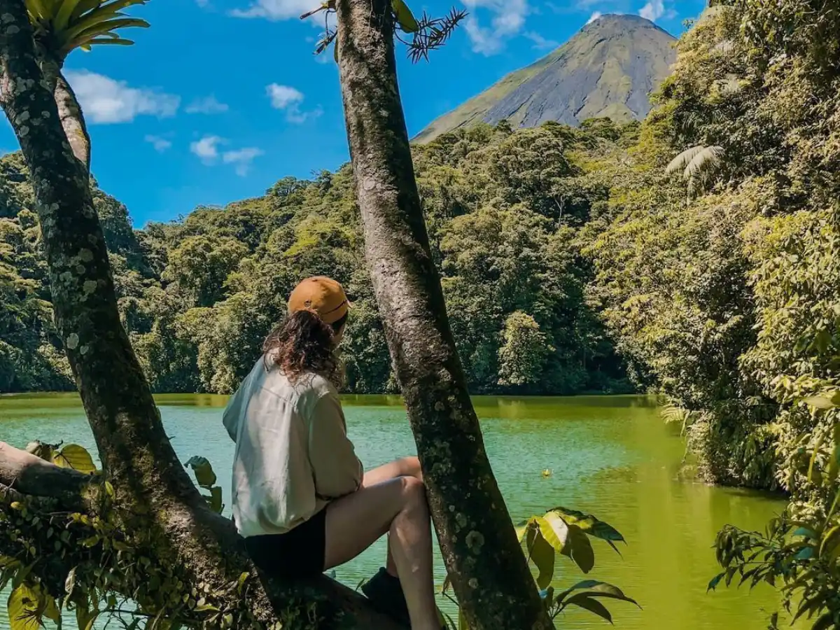 Persona sentada en la horqueta de un árbol sobre una laguna verde, con el volcán a lo lejos