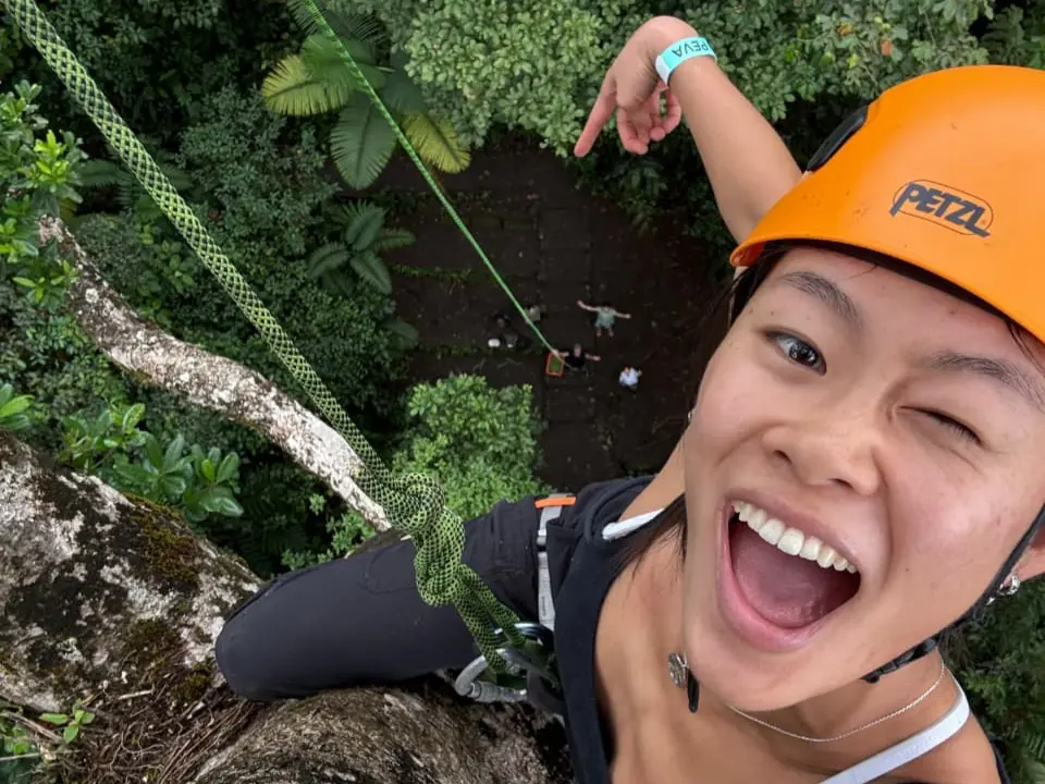 Smiling climber in an orange helmet taking a selfie high on a tree, with the forest floor far below