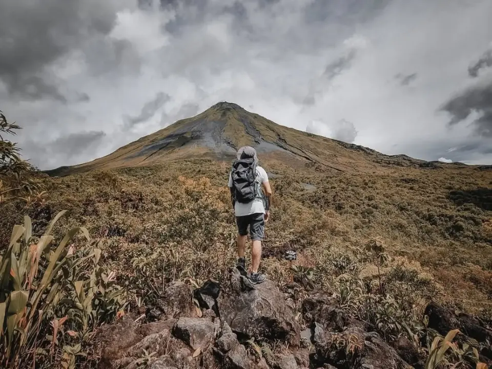 Hiker with a backpack walking up a rocky slope toward a volcano wrapped in clouds