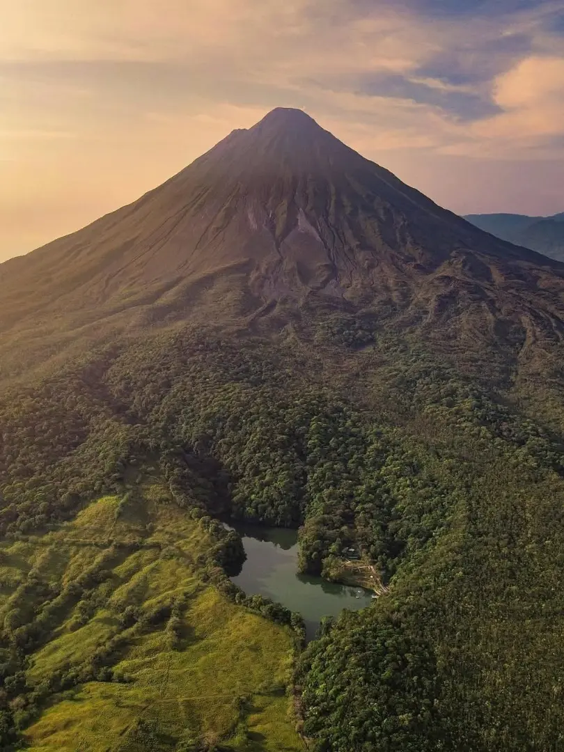 Vista aérea del Volcán Arenal sobre el bosque denso al atardecer, con una pequeña laguna y pasto verde abajo