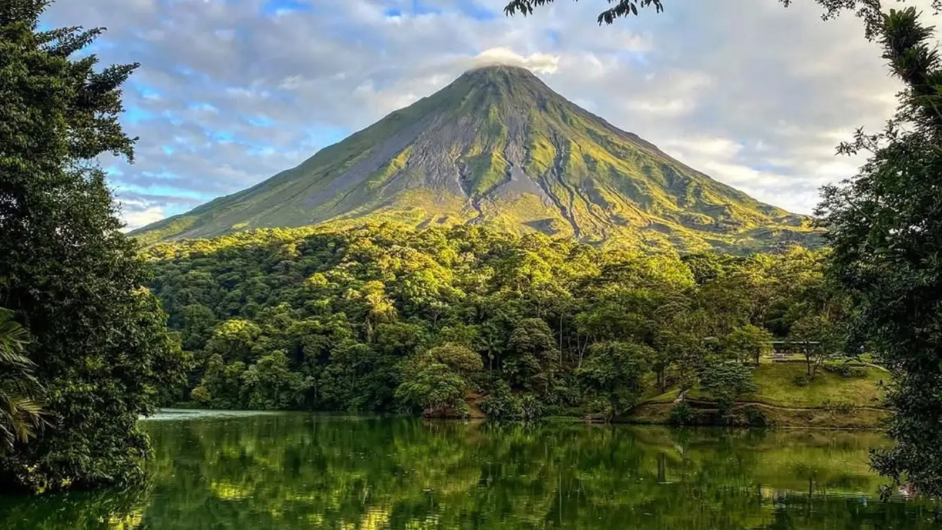 Arenal Volcano in warm light, framed by trees and reflected in a still green lake
