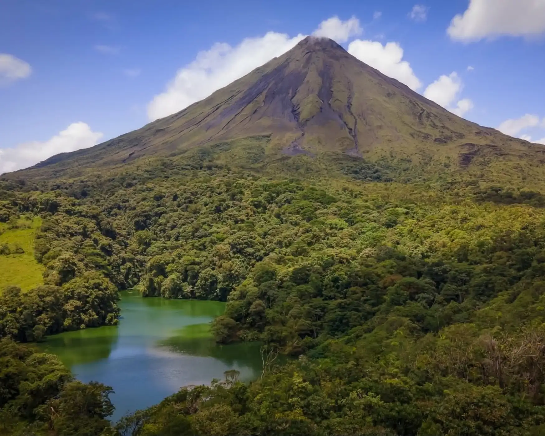 Arenal Volcano with a cloud at its summit above dense green forest and a small green lagoon
