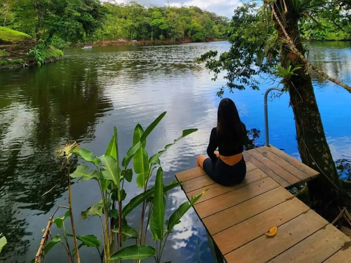 Woman sitting on a wooden dock beside a calm lagoon surrounded by forest