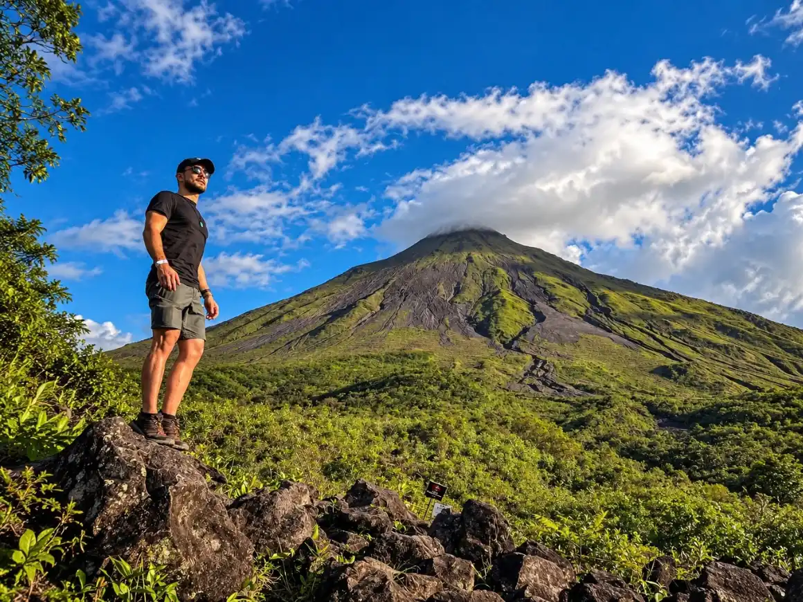 Hiker standing on dark lava rocks with Arenal Volcano and a blue sky behind