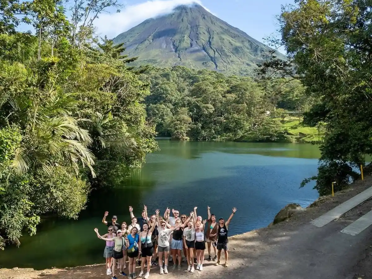 Group of hikers raising their arms beside a green lagoon, with Arenal Volcano behind the forest