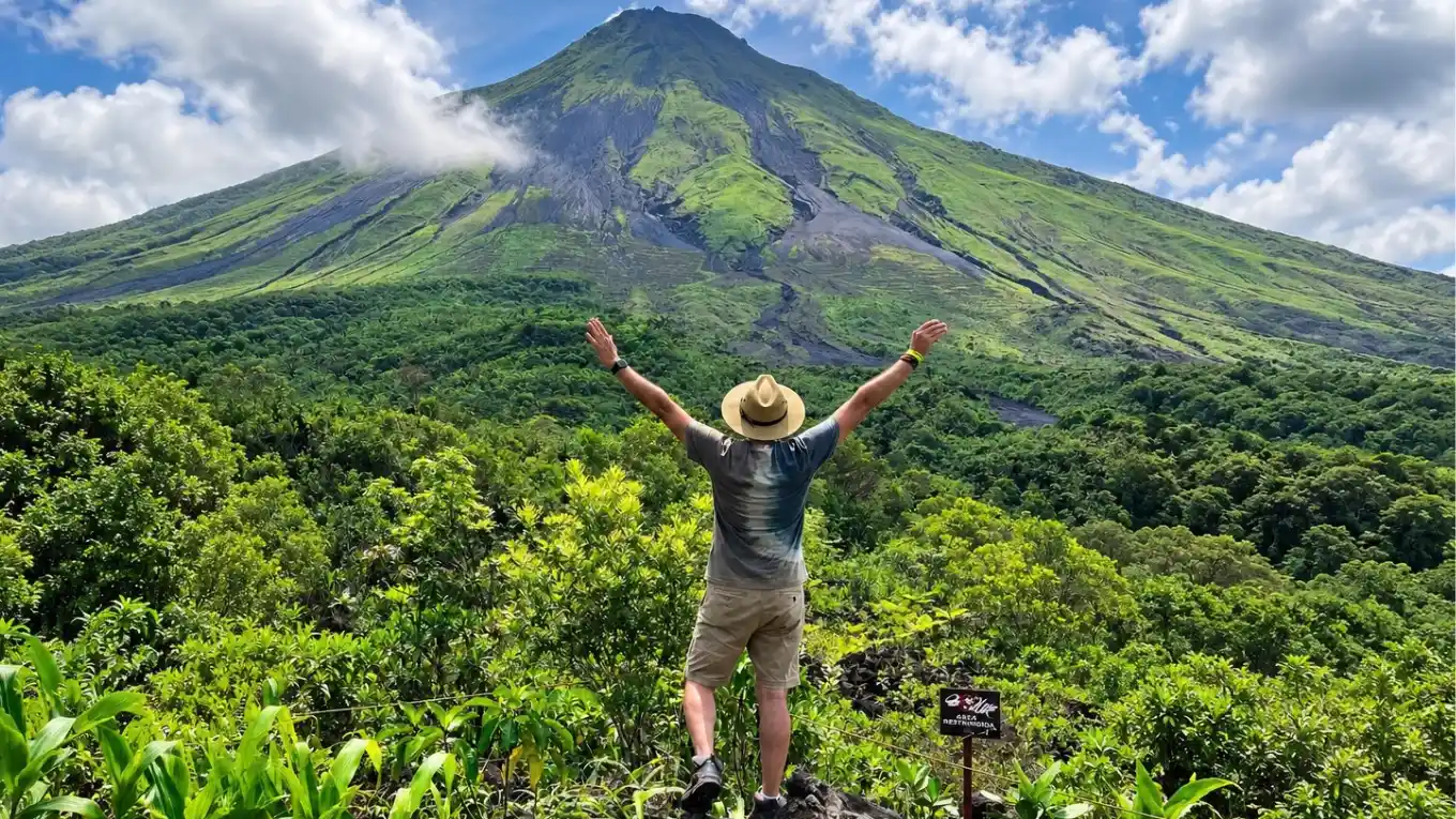 Hiker in a straw hat with raised arms facing the green slopes of Arenal Volcano